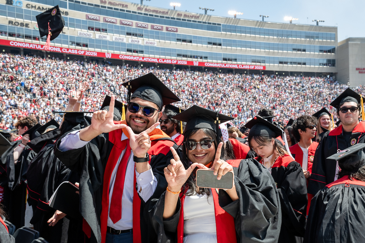 Students in graduation caps and gowns smiling and making a W with their fingers at a graduation ceremony with many other students in the background