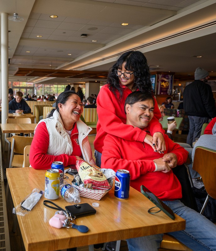 Two adults sitting at a table, one adult standing with their arms around one of the adults at the table, all smiling.