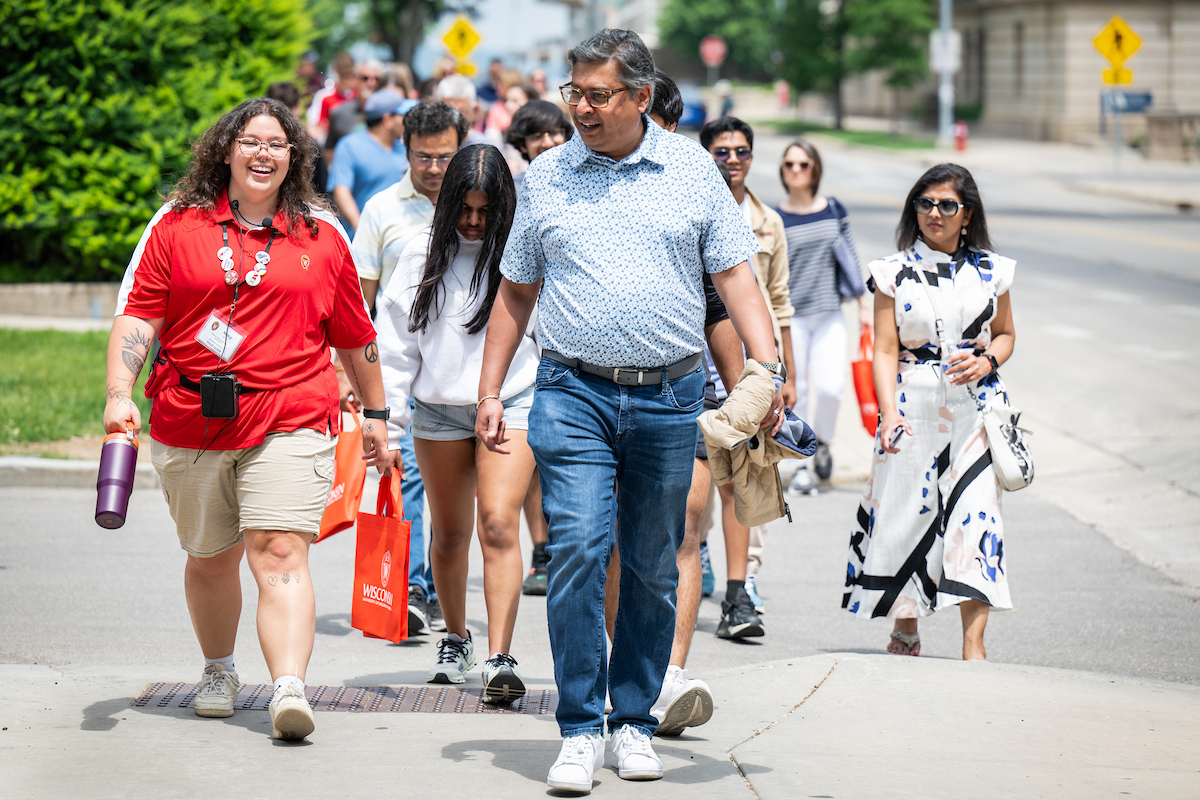 A tour guide wearing a red UW-Madison shirt walks with a group of students and parents down a street.