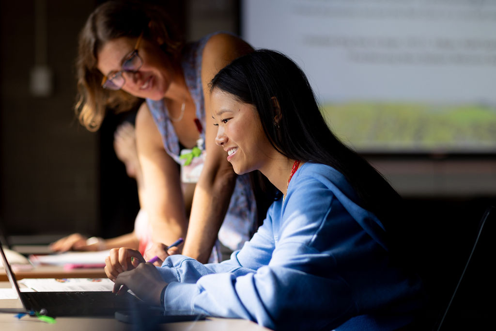 Advisor leaning toward a student who is seated and looking at a laptop
